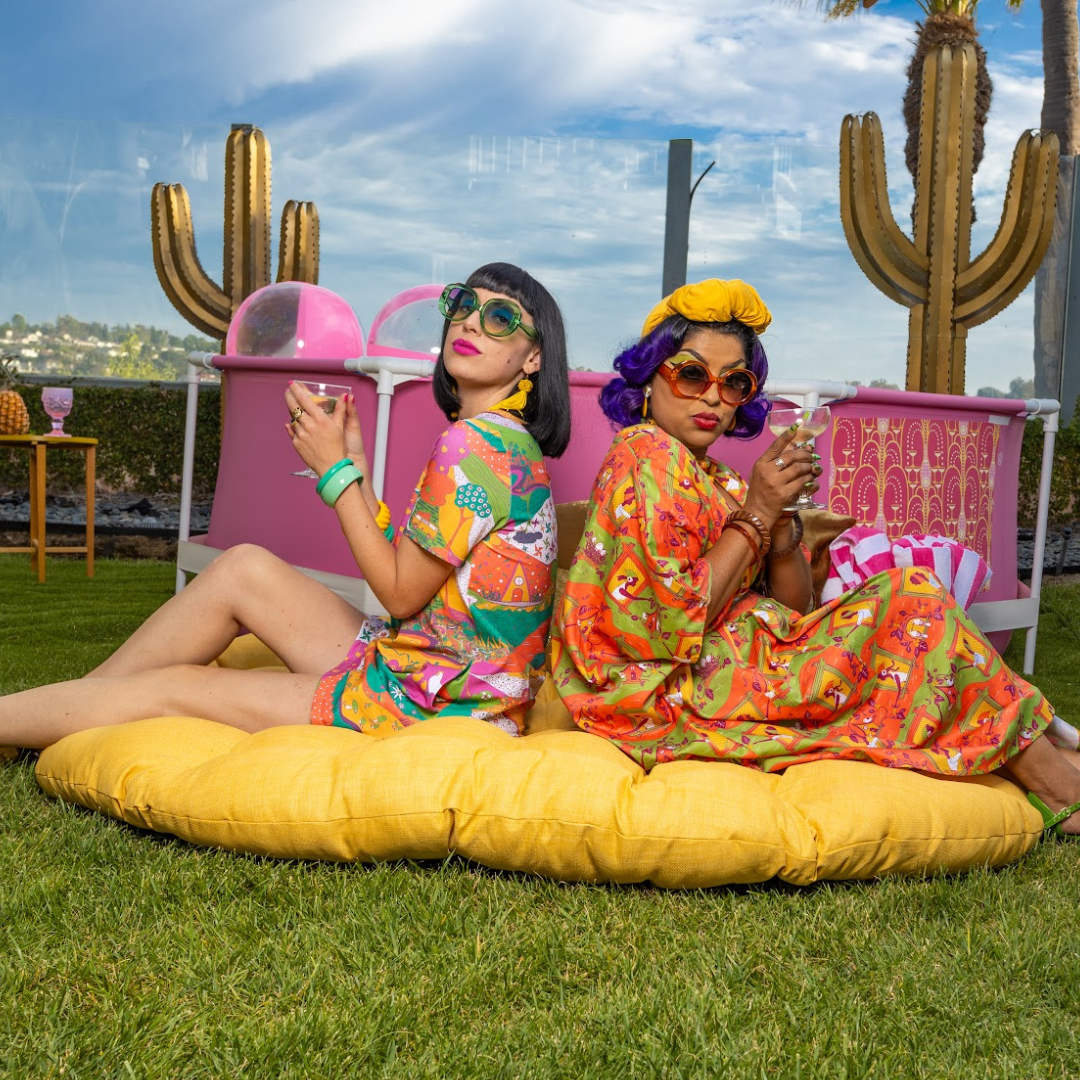 Model in short pocket tunic sitting with model in colorful caftan on cushion