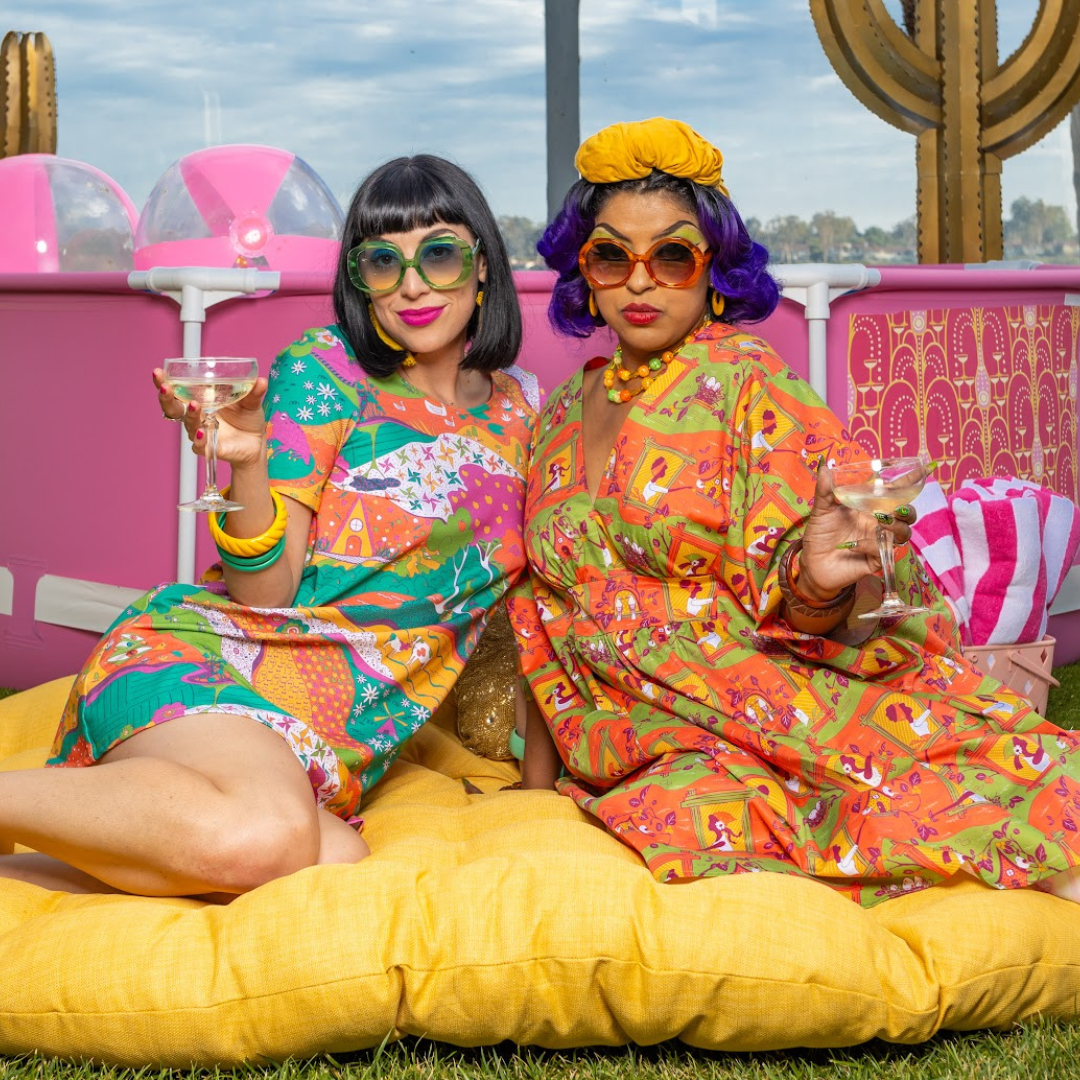 Black-haired model in short pocket tunic sitting with model in colorful caftan