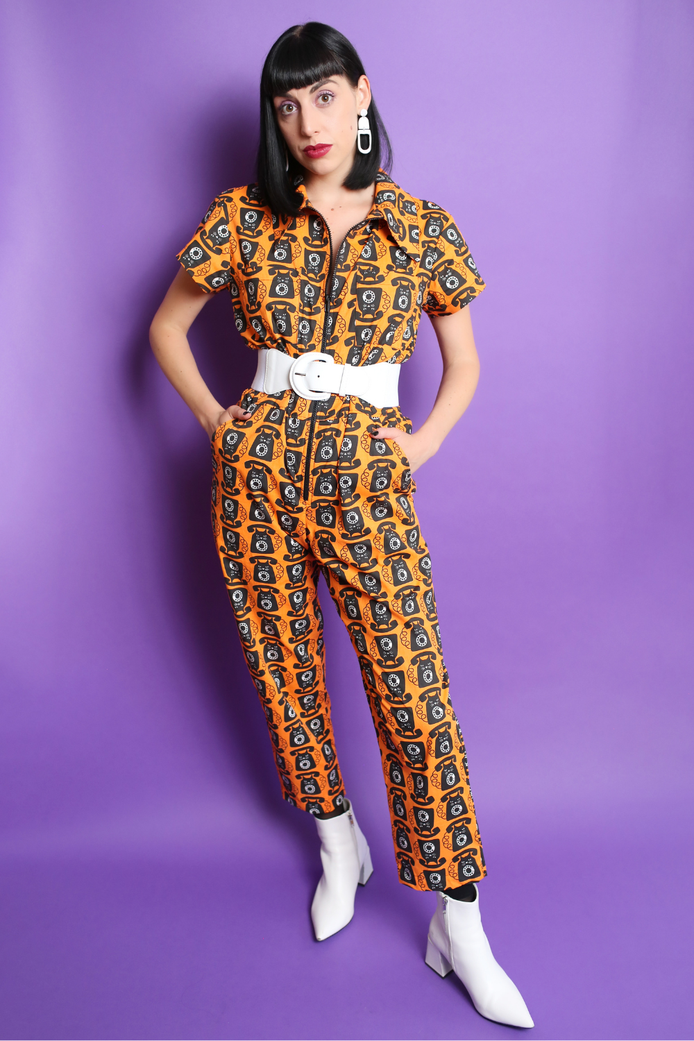Black-haired model in orange and black cat phone print jumpsuit with white accessories
