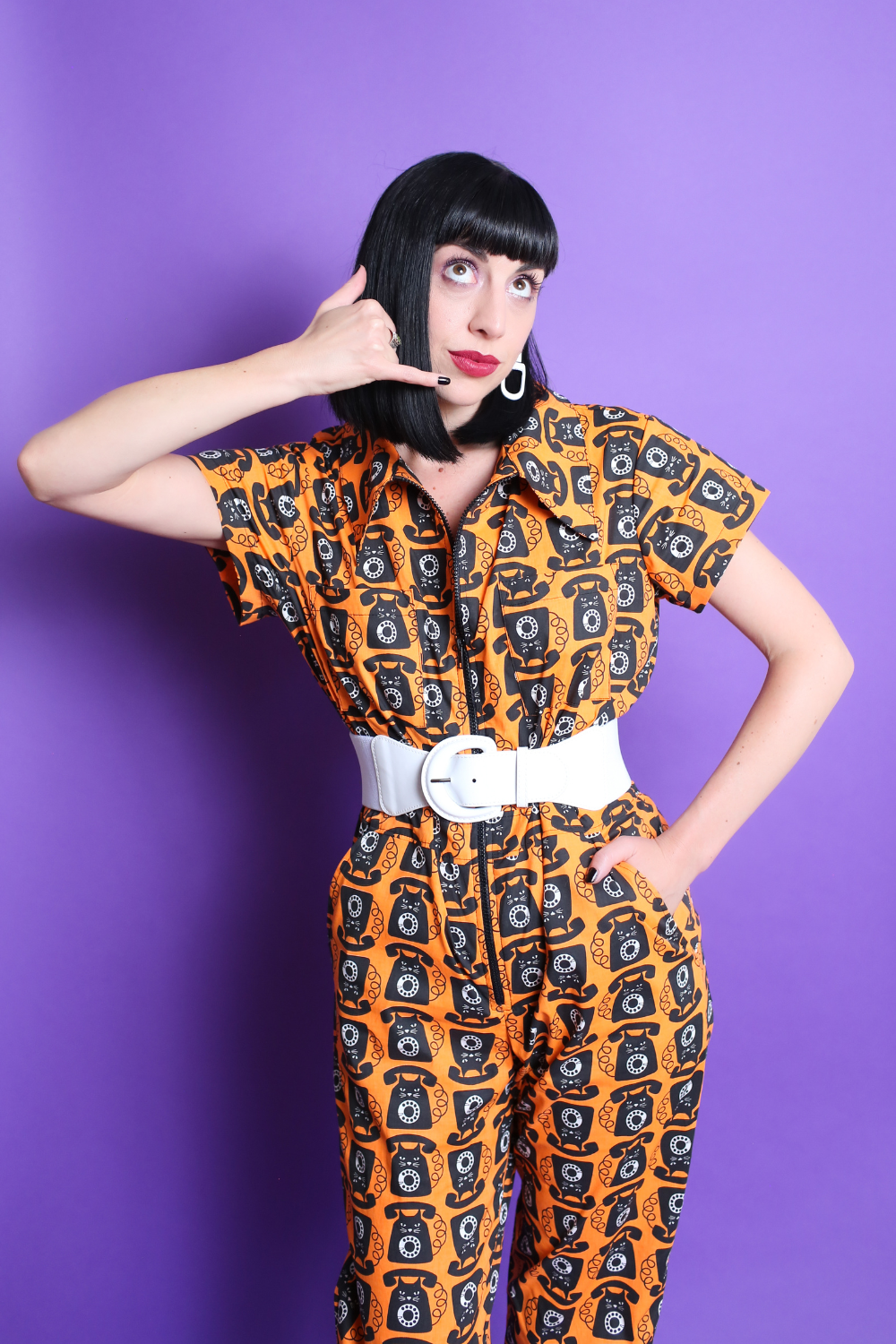 Black-haired model in orange and black cat phone print jumpsuit with white accessories