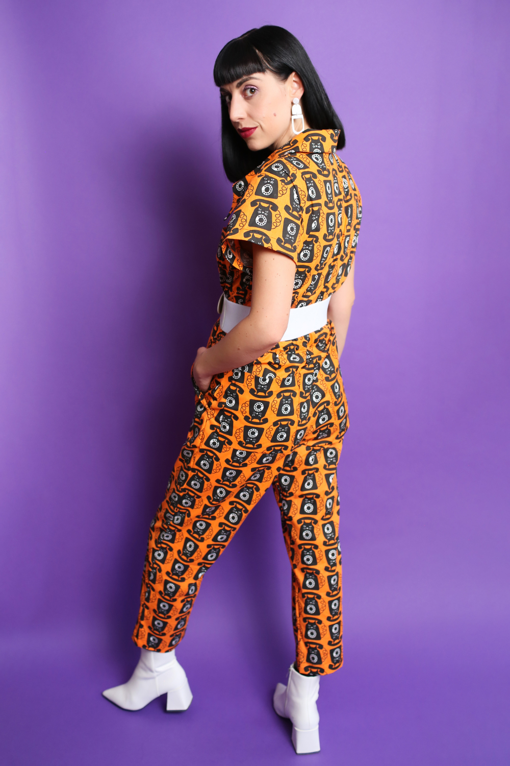 Black-haired model in orange and black cat phone print jumpsuit with white accessories