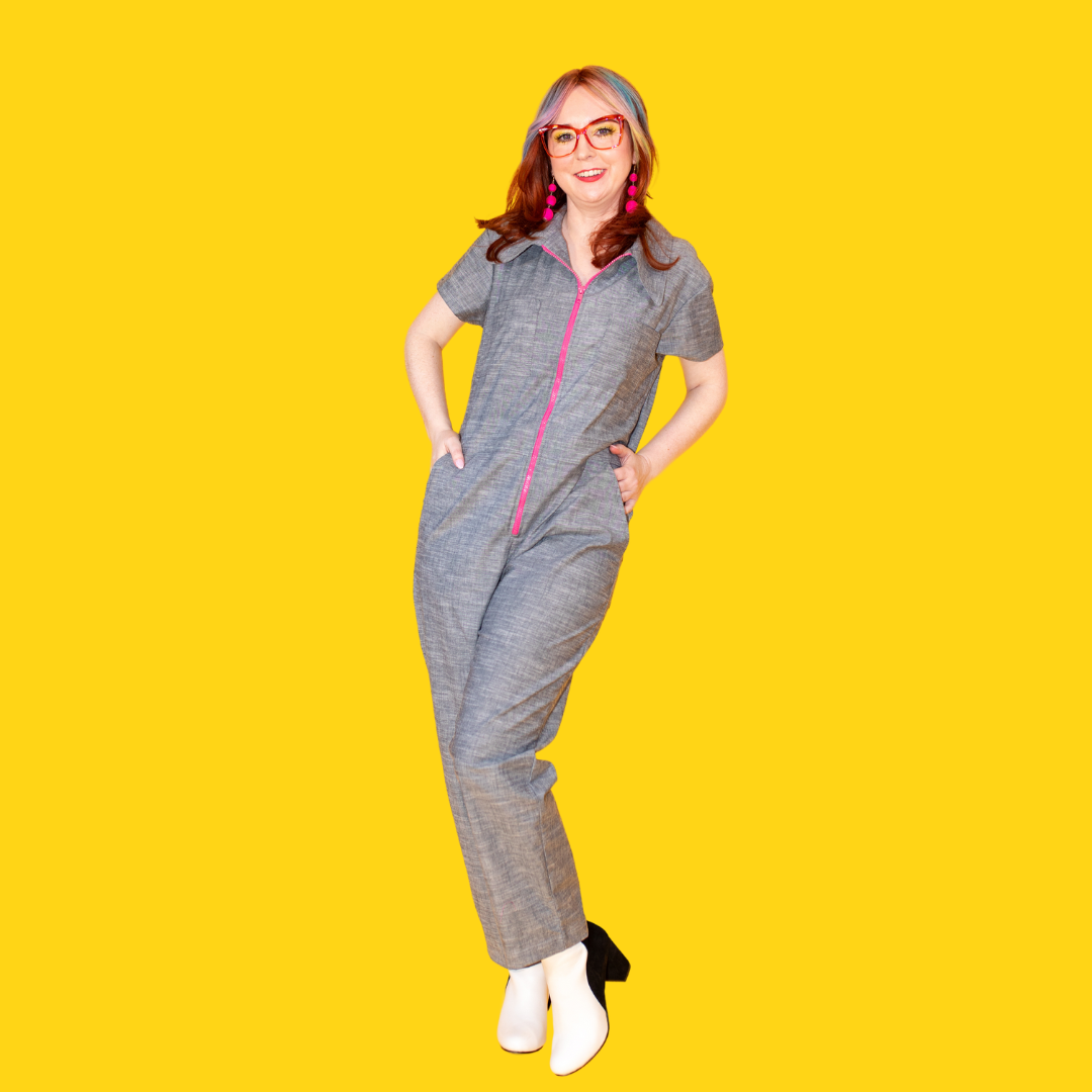 Model wearing glasses and greyish blue champbray jumpsuit with bright pink zip