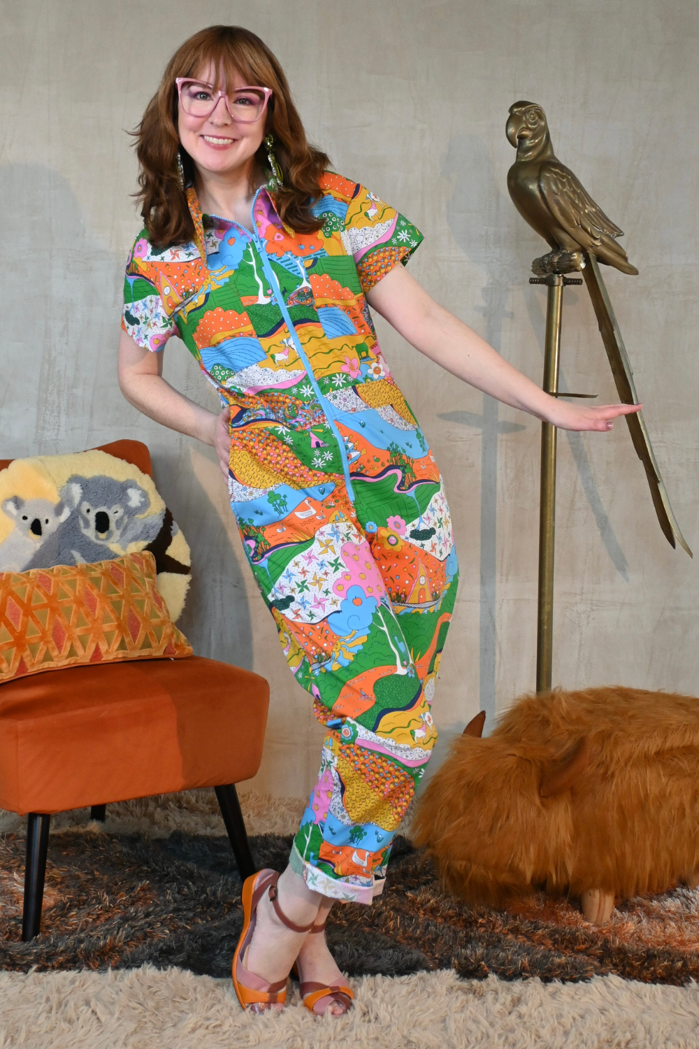 Girl with glasses wearing rainbow print jumpsuit, standing next to metal parrot