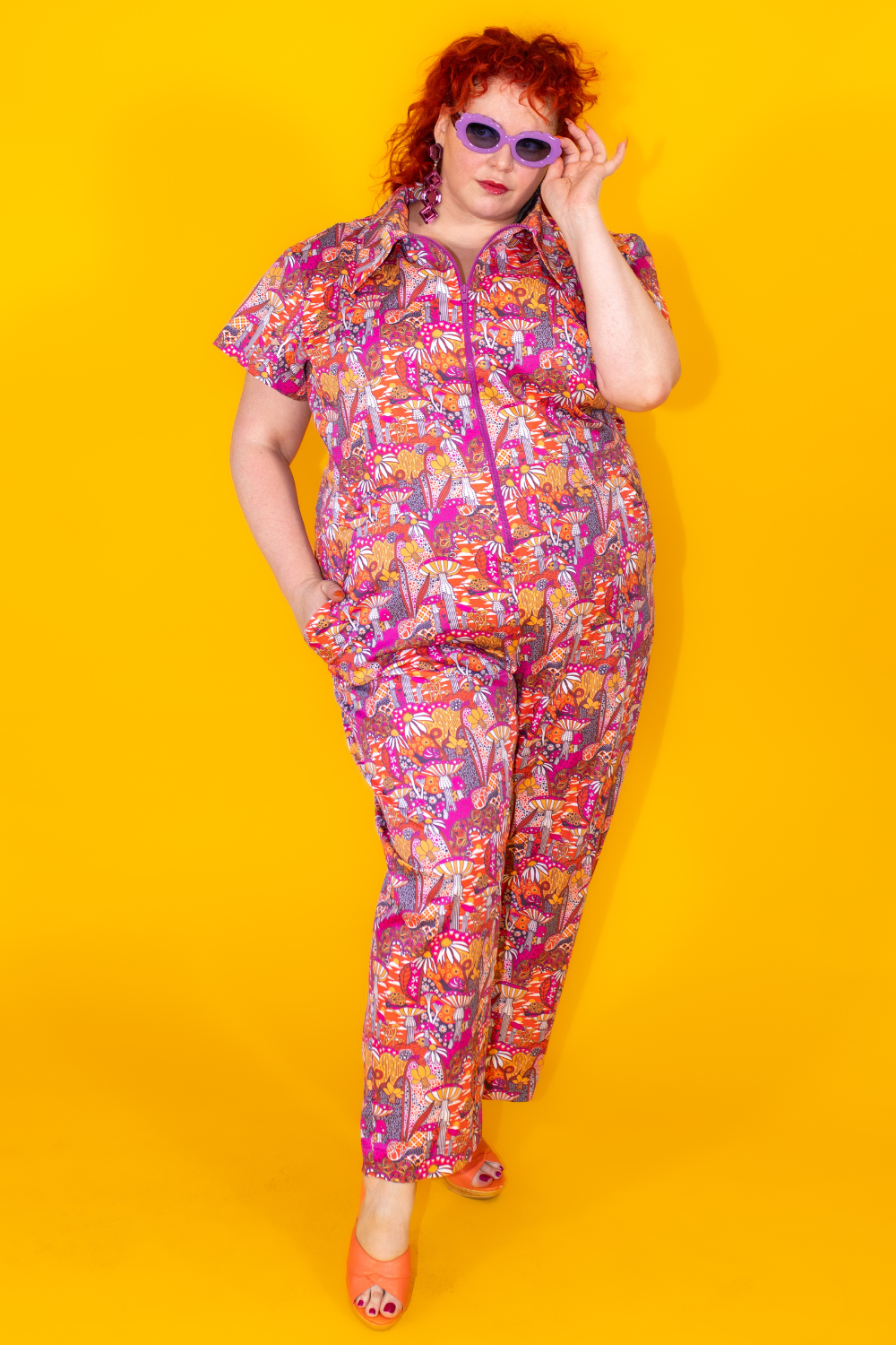 Red-haired model in bright mushroom print jumpsuit and purple sunglasses