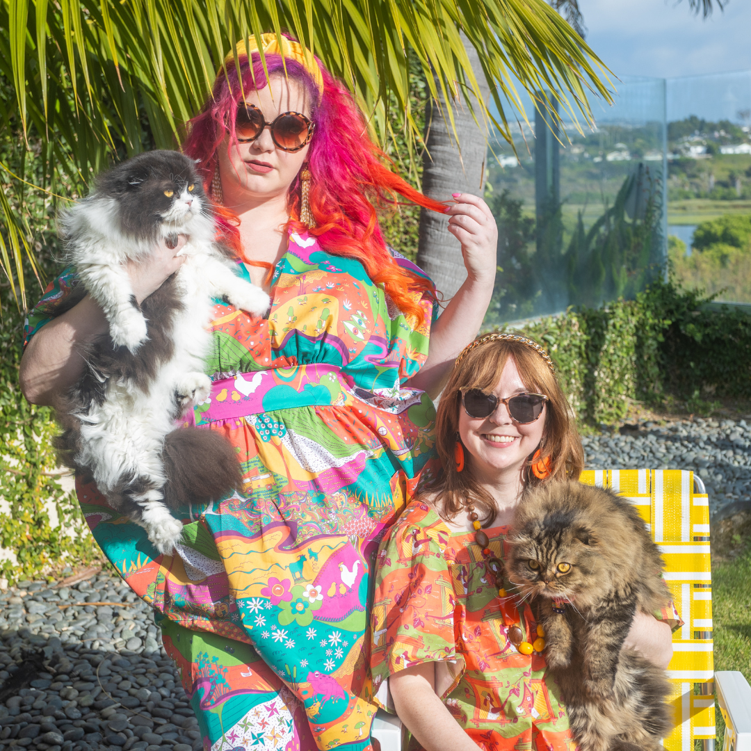 Model in colorful caftan and model in colorful dress, both holding cats