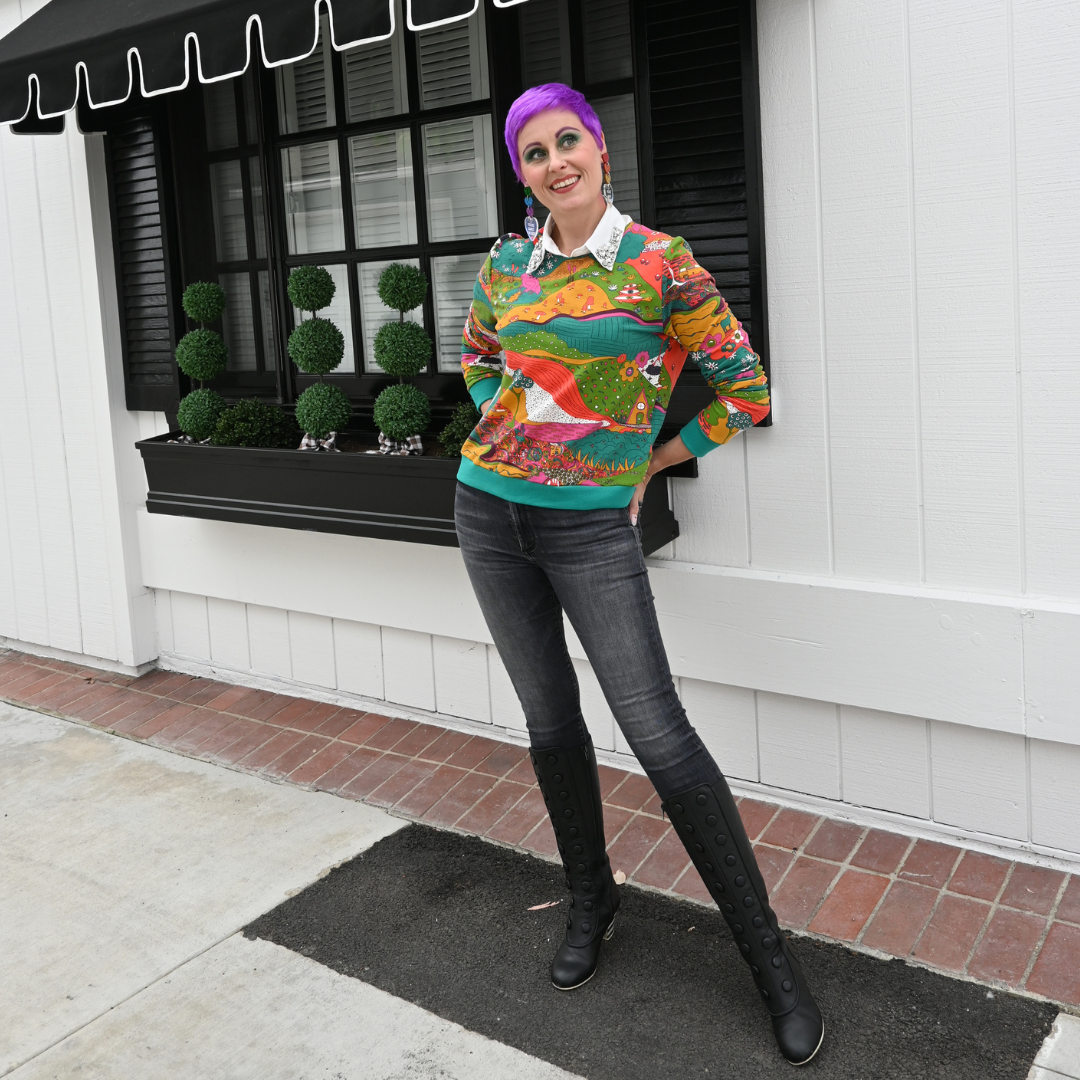Full body view of purple-haired model wearing landscape print sweatshirt and standing in front of a window