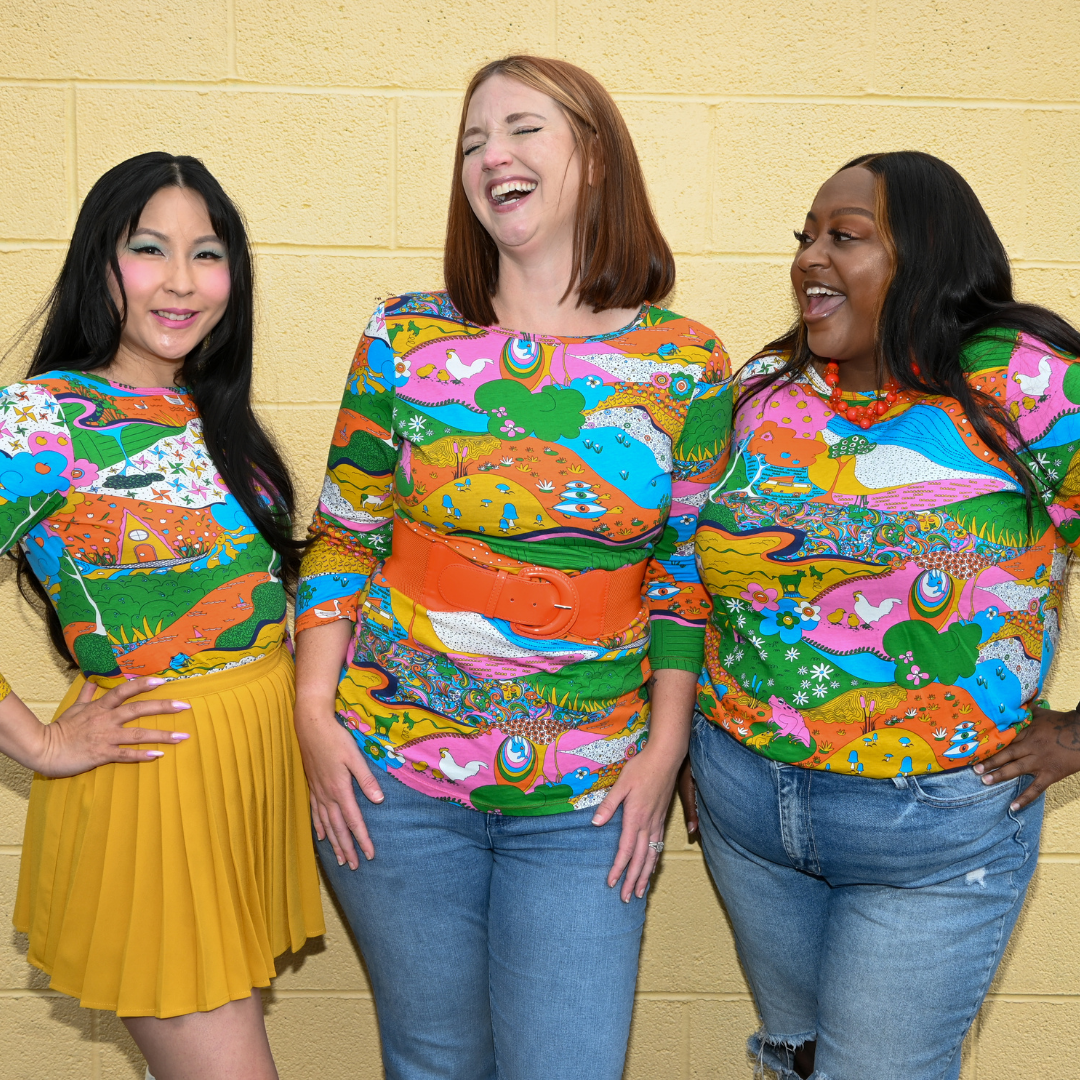 Closeup of 3 smiling models in matching landscape print tees featuring colorful animals and natural elements