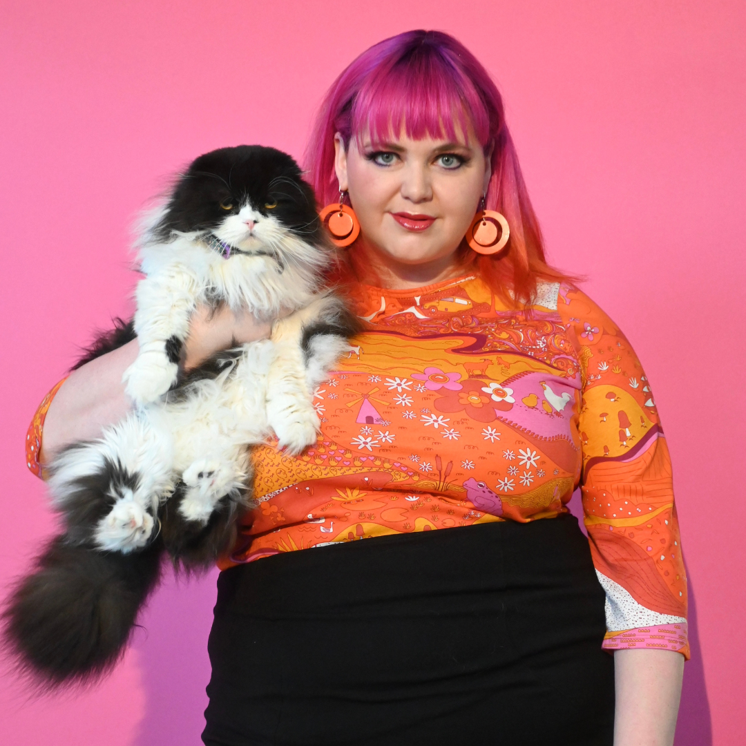 Pink-haired girl holding cat and wearing orange pink red 3/4 graphic tee