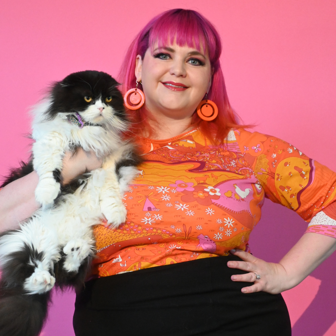 Cute pink-haired girl in fun earrings and wearing orange pink red landscape print graphic tee and holding cat