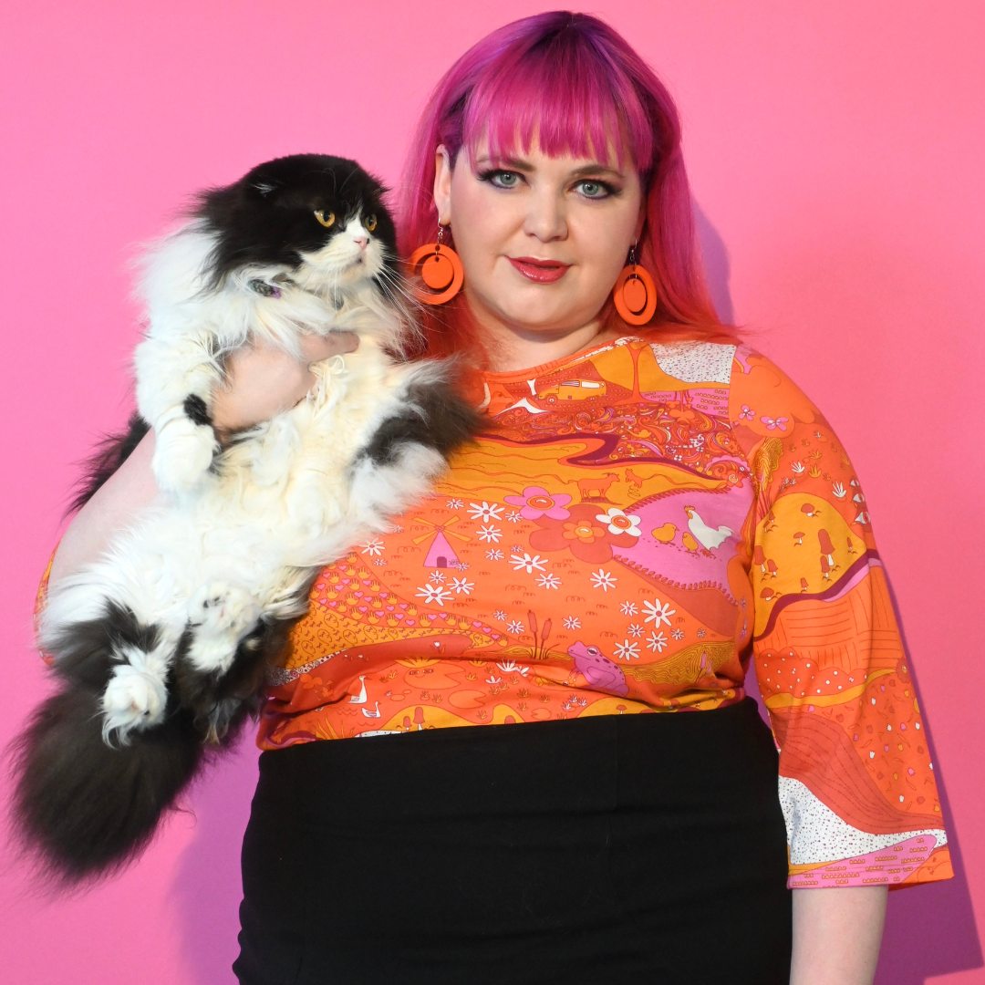 Cute pink-haired girl holding cat and wearing bright orange pink red landscape print graphic tee