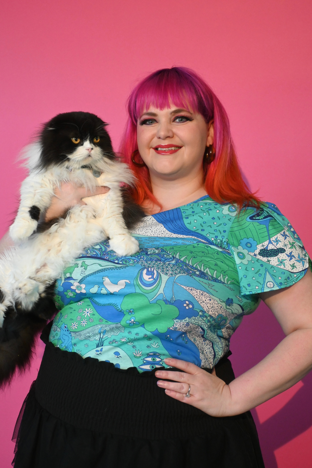 Pink haired model holding cat wearing shirt with landscape graphic in green and blue