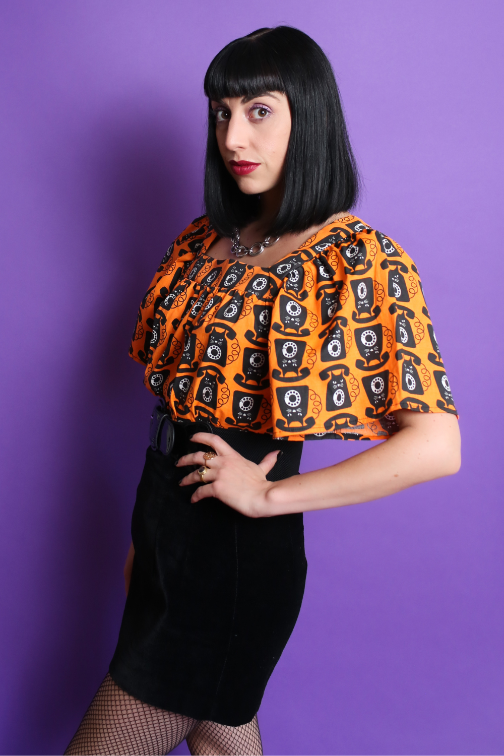 Black haired model in orange and black cat phone print top & mini skirt, side view