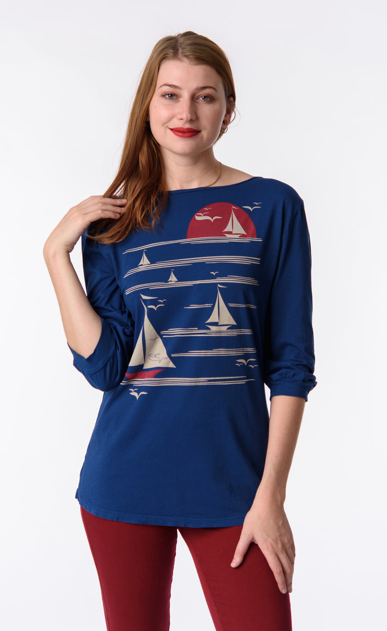 Blonde model wearing long sleeve blue shirt with graphic of boats on the water at sunset