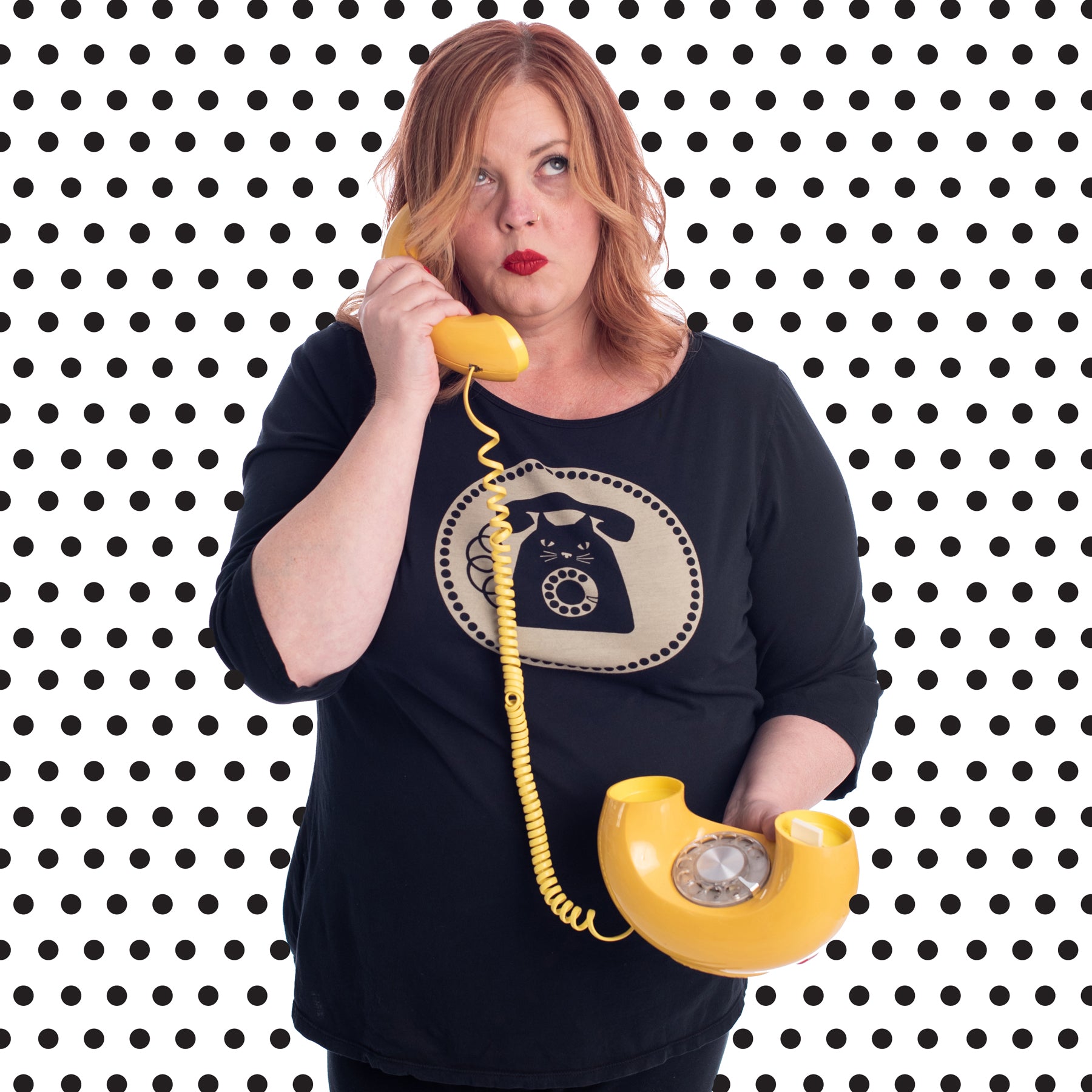 Red haired plus size model talking on donut phone while wearing black cat phone graphic 3/4 sleeve tee and with polka dot background