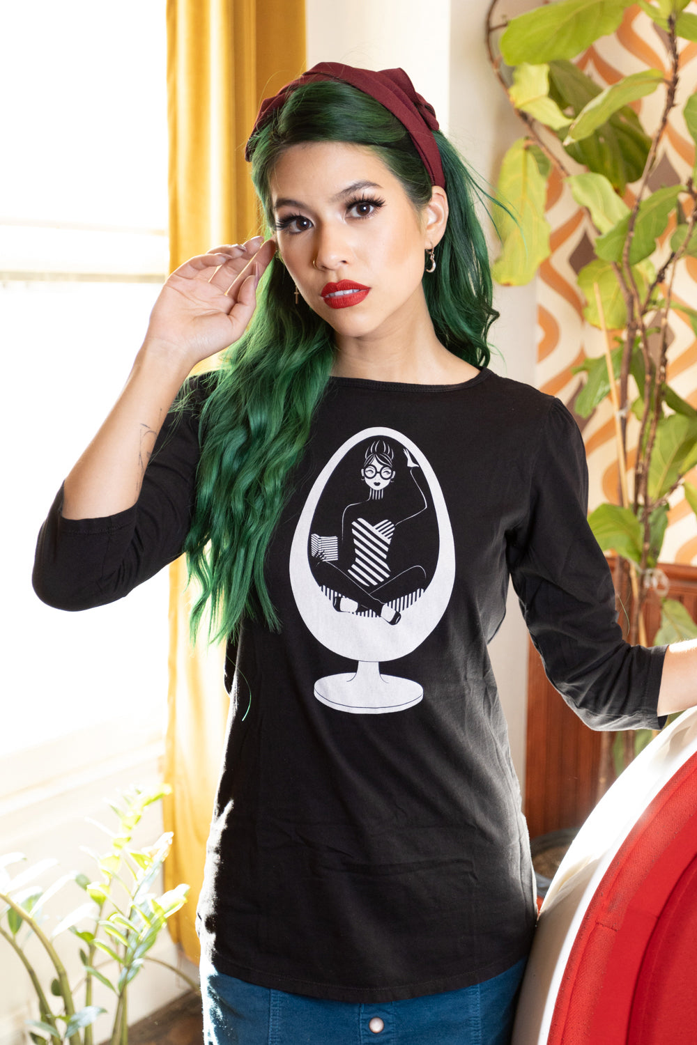 Girl with green hair standing next to ball chair and wearing black 3/4 sleeve tee with a white graphic of a girl reading in an egg chair