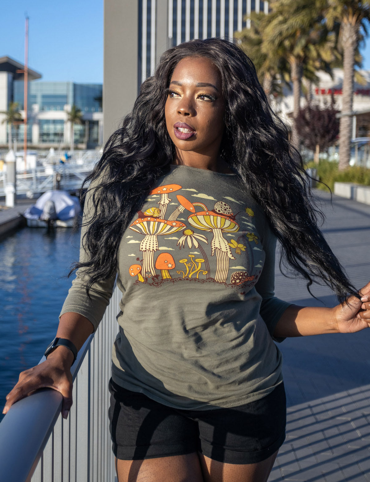 Long-haired model in olive green 3/4 sleeve tee with mushrooms, snails and flowers, standing at railing