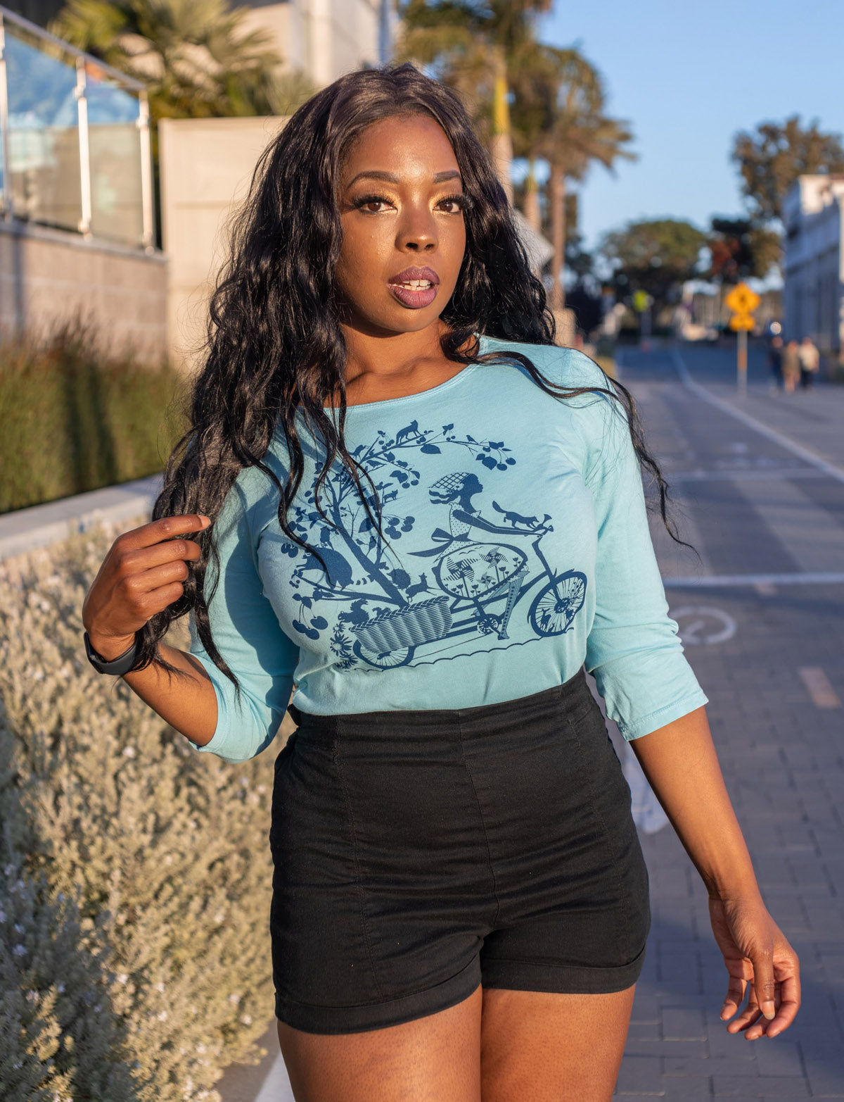 Long-haired model in blue 3/4 sleeve tee with print of girl riding bicycle with fruit trees and animals