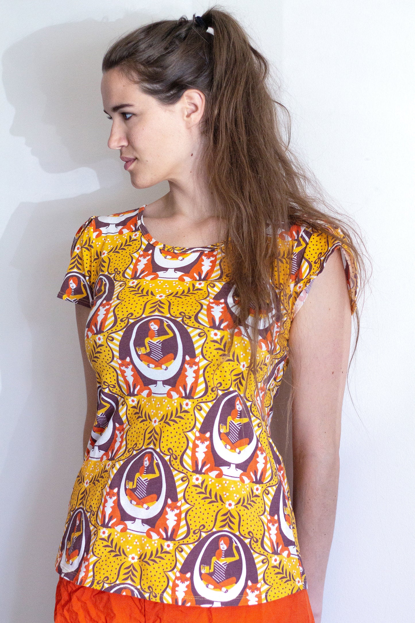 Model in yellow tulip-sleeve tee with foxes, girl reading in egg chair print in orange, brown and white