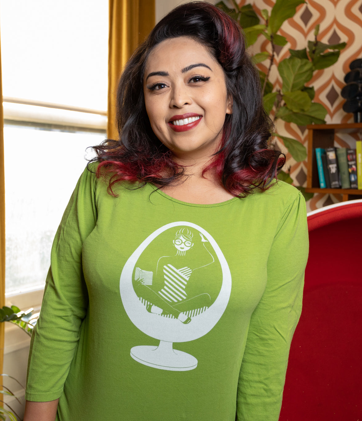 Model in apple green 3/4 sleeve tee with vintage-inspired print of girl with glasses reading in an egg chair