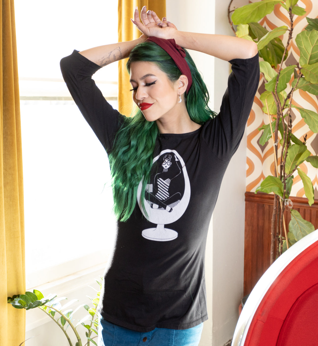 Long-haired girl standing in a sunny window and wearing black tee with white print of girl in a vintage chair