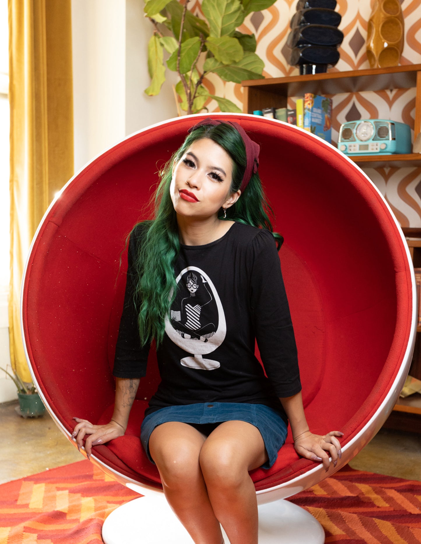 Long-haired girl sitting in ball chair wearing black 3/4 sleeve tee with white graphic of girl reading in a vintage egg chair