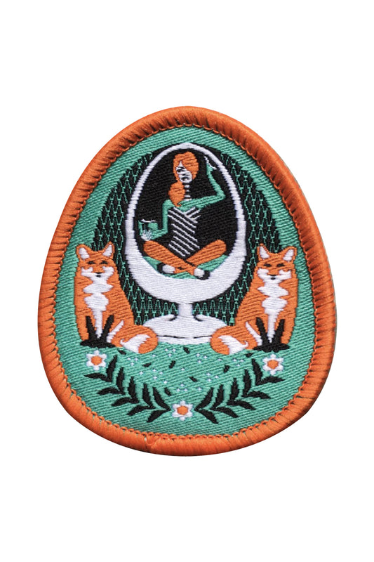 Green and orange iron-on patch featuring a girl in an egg chair with 2 foxes and flower accents