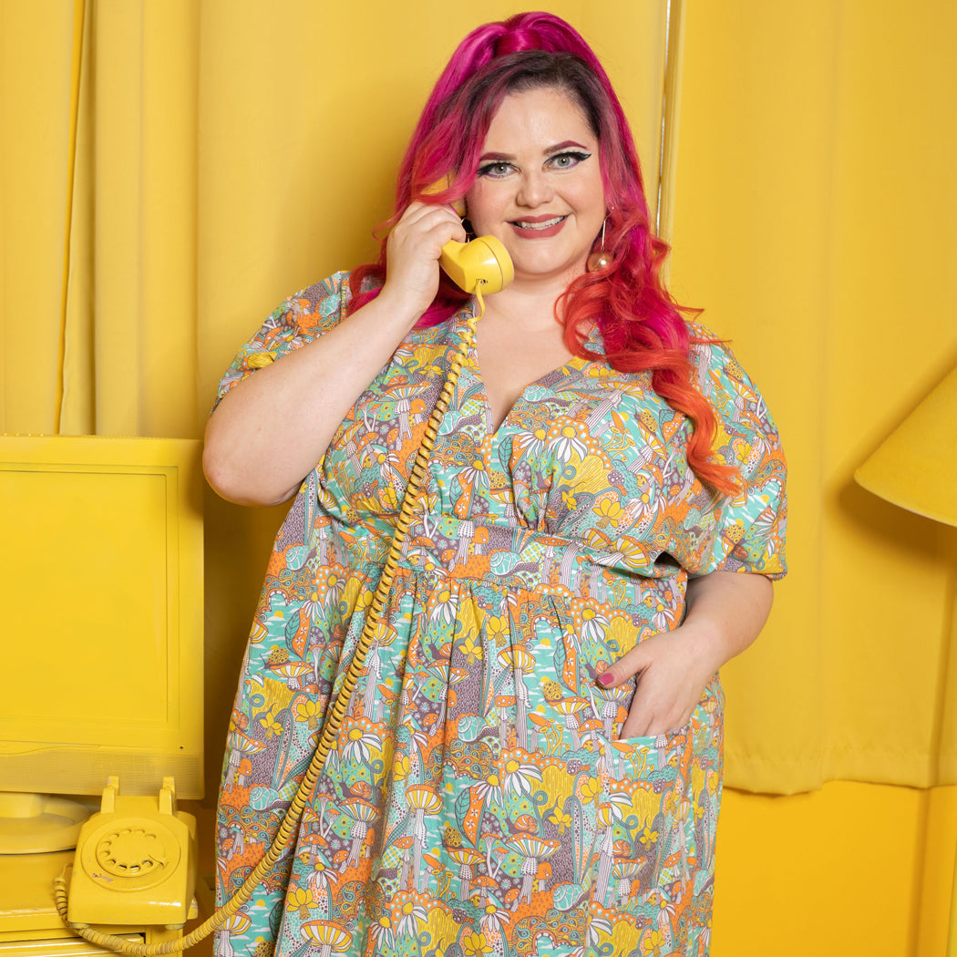 Pink haired model in bright green, yellow and orange mushroom, snail and flower print v-neck below knee caftan dress in yellow room