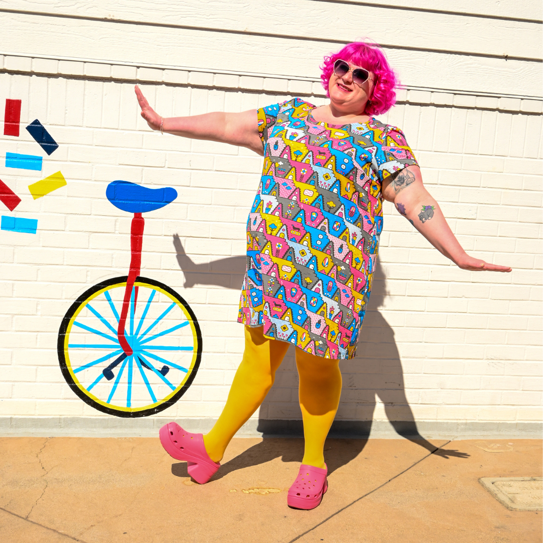 Pink-haired model wearing darling a-frame tunic in front of wall with unicycle design