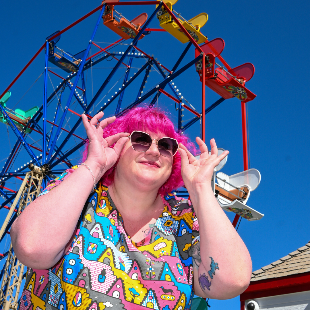 Closeup of pink-haired model in geometric print tunic and fun sunglasses with ferris wheel in the background
