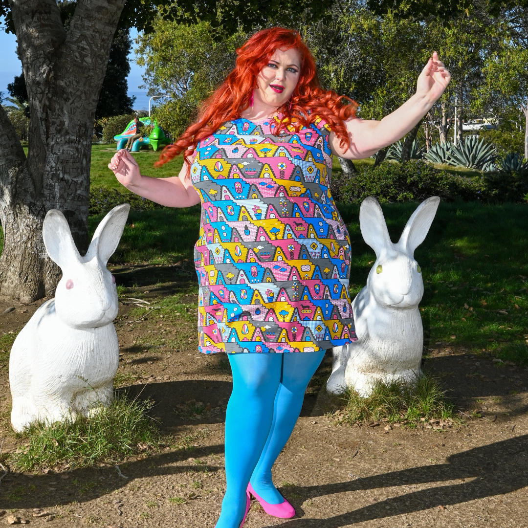 Gorgeous red-haired model posing in pastel colored geometric tunic in pink, yellow and blue, surrounded by bunny statues