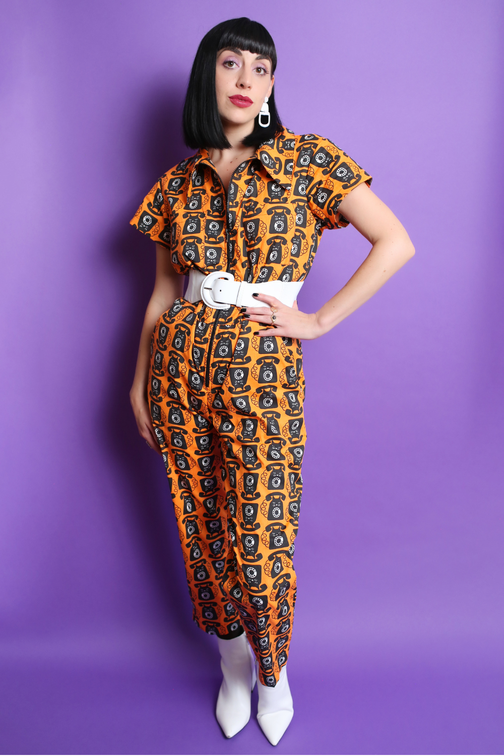 Black-haired model in orange and black cat phone print jumpsuit with white accessories