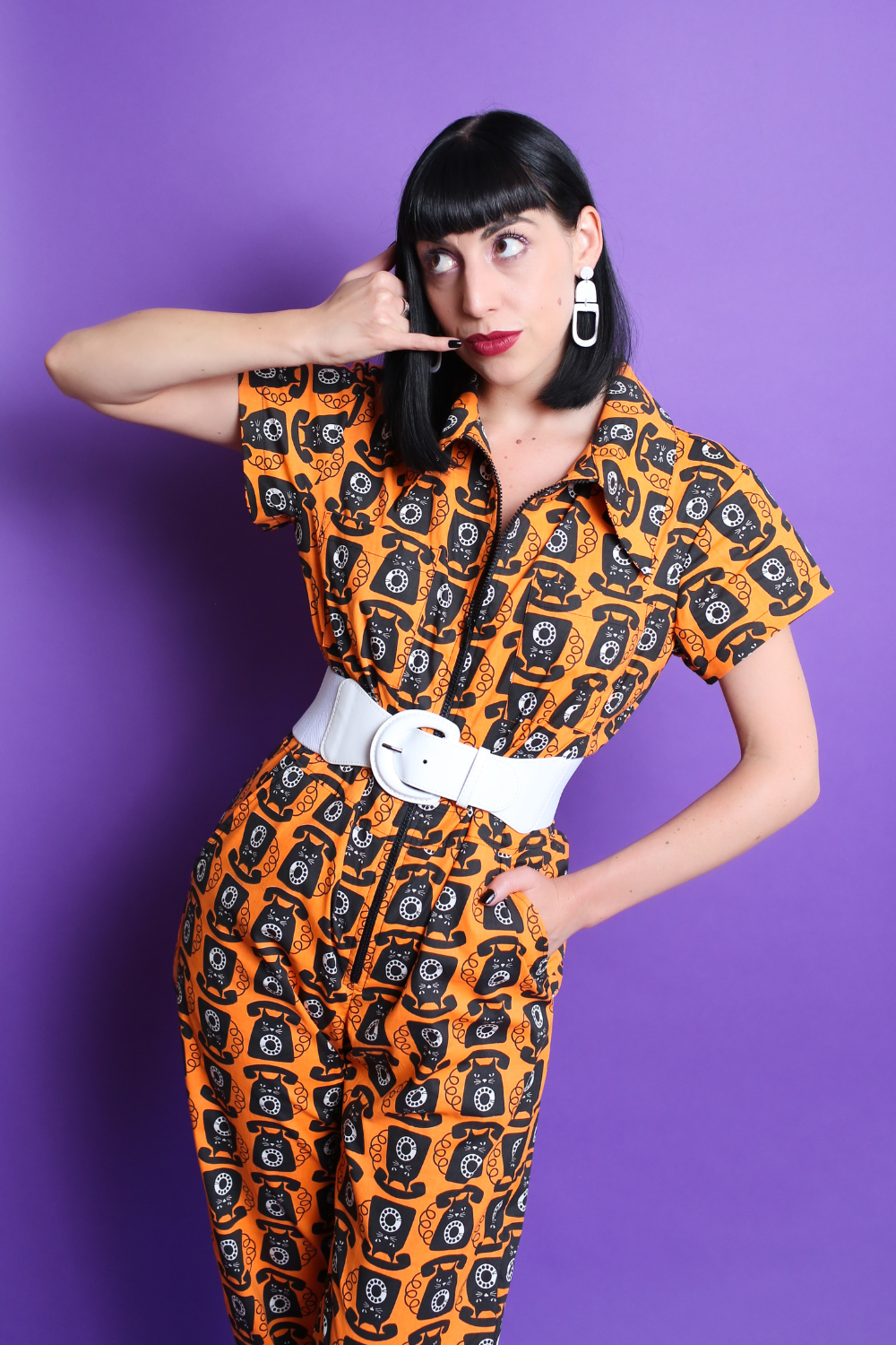 Black-haired model in orange and black cat phone print jumpsuit with white accessories