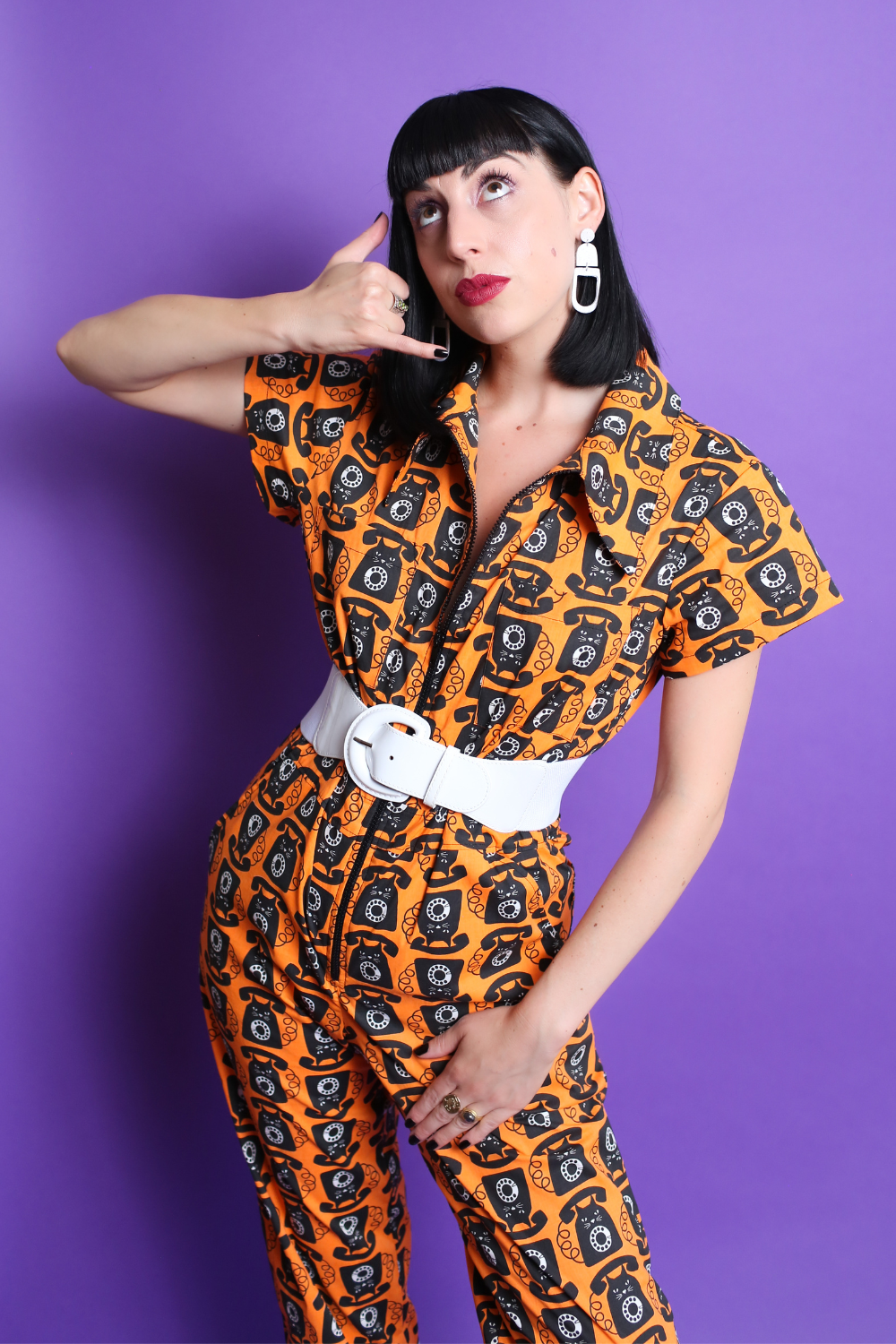Black-haired model in orange and black cat phone print jumpsuit with white accessories