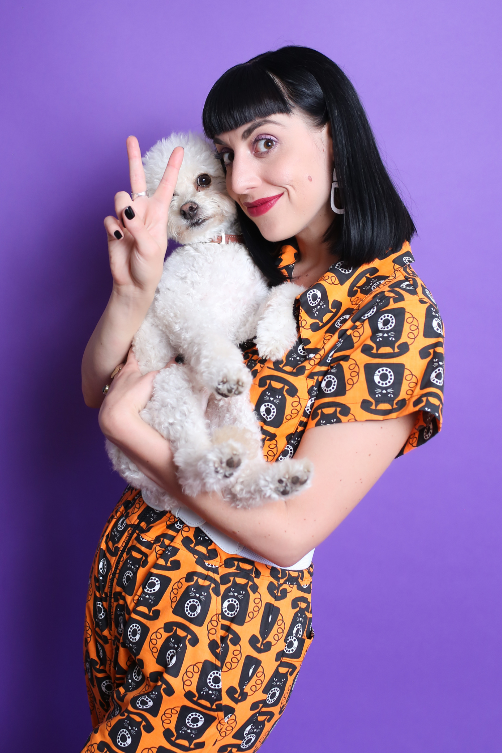 Black-haired model in orange and black cat phone print jumpsuit with white accessories and a small white dog