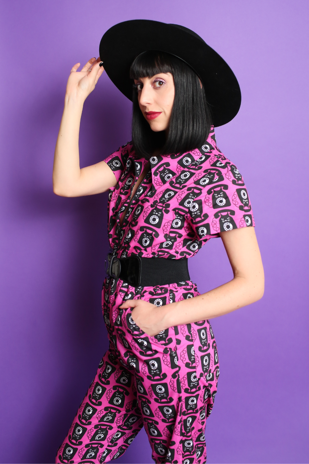 Black haired model in black hat and pink and black printed jumpsuit, side view