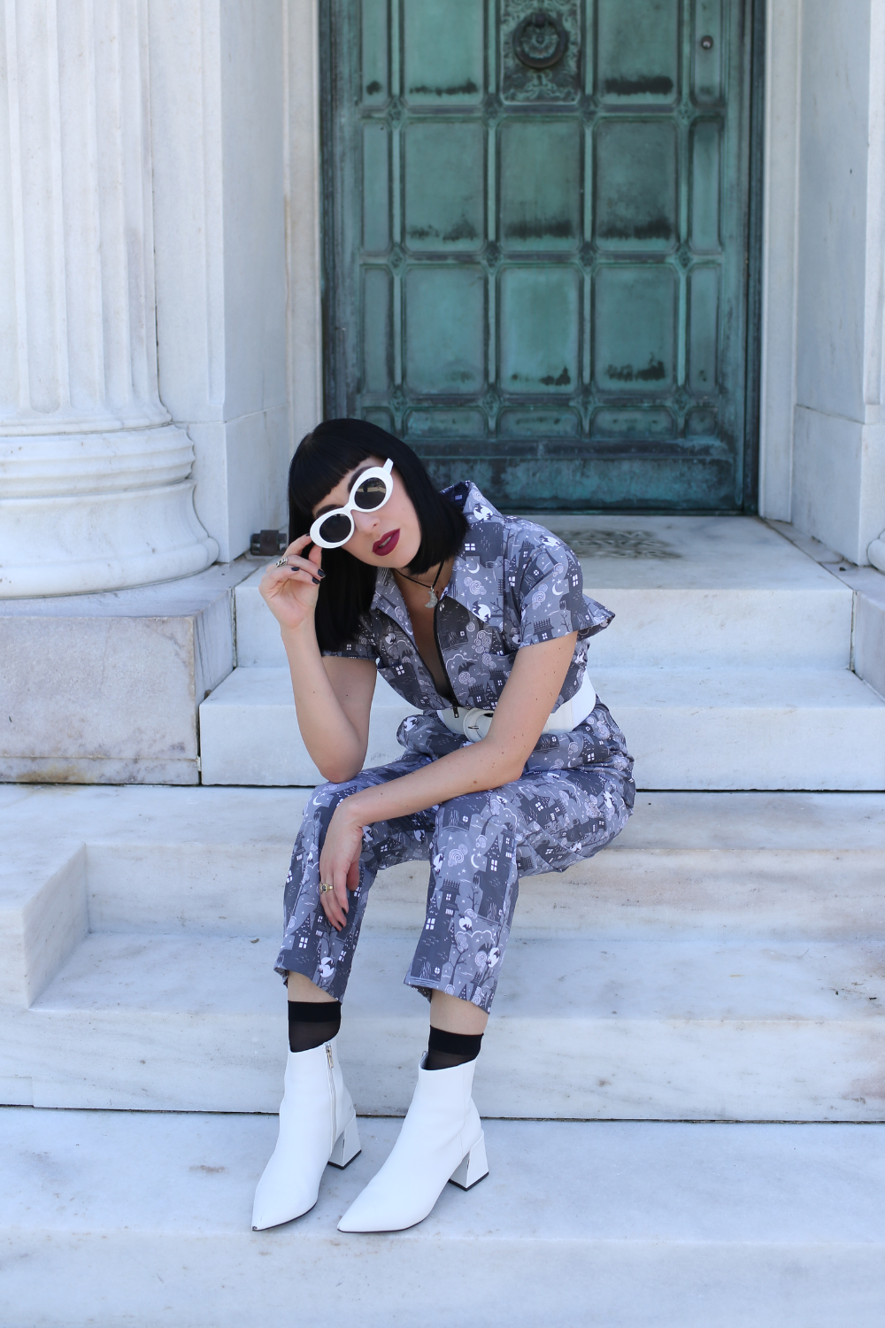 Tall model wearing grey haunted house print jumpsuit , sitting on steps, with white accessories