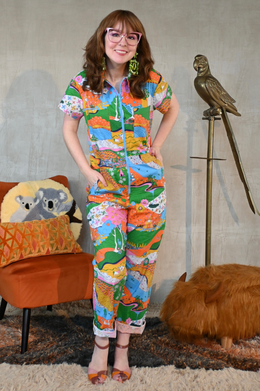 Girl with glasses wearing rainbow print jumpsuit, standing next to metal parrot