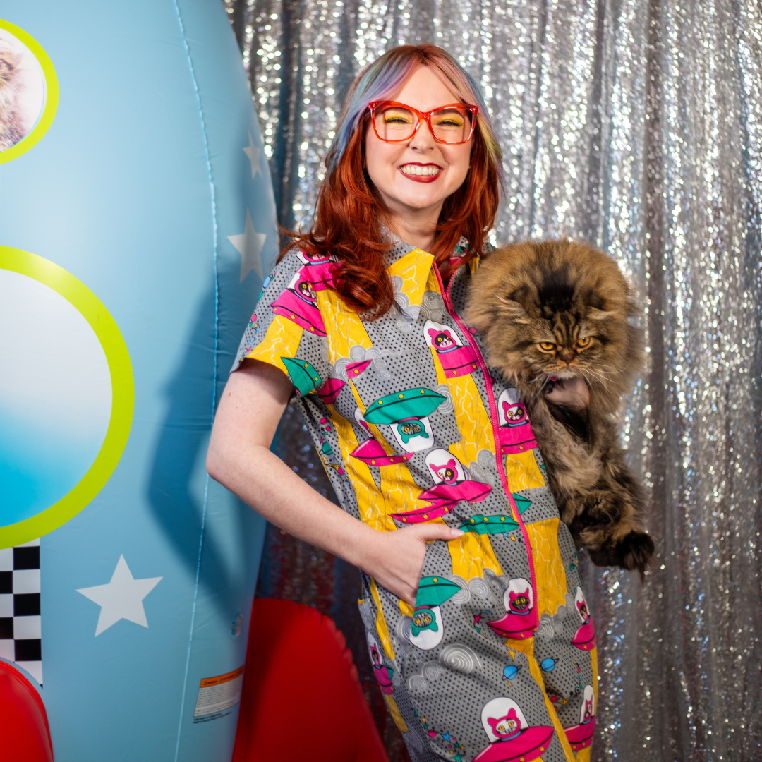 Closeup of model in classes holding a cat and wearing a jumpsuit with a print of cats in space ships