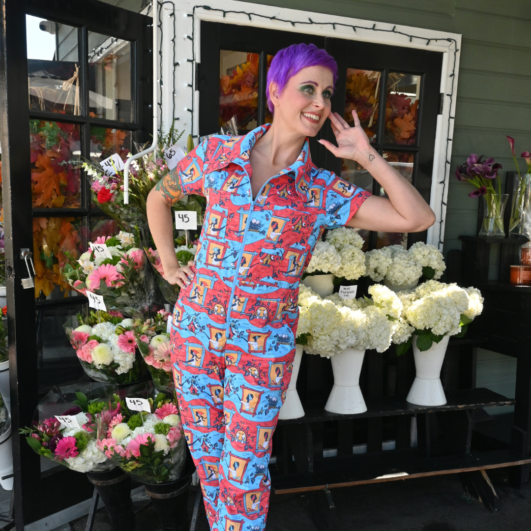 Purple-haired model in red, yellow and blue jumpsuit with girls in windows print posing in front of flower shop