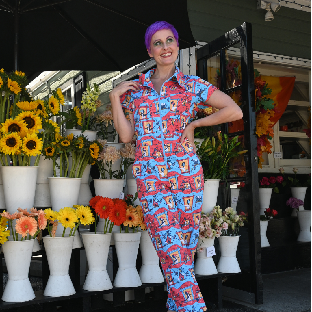 Purple-haired model in red, yellow and blue jumpsuit with girls in windows print surrounded by flowers