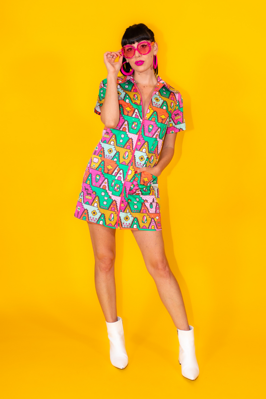 Dark-haired model in bright zip front geometric minidress with white ankle boots and pink sunglasses