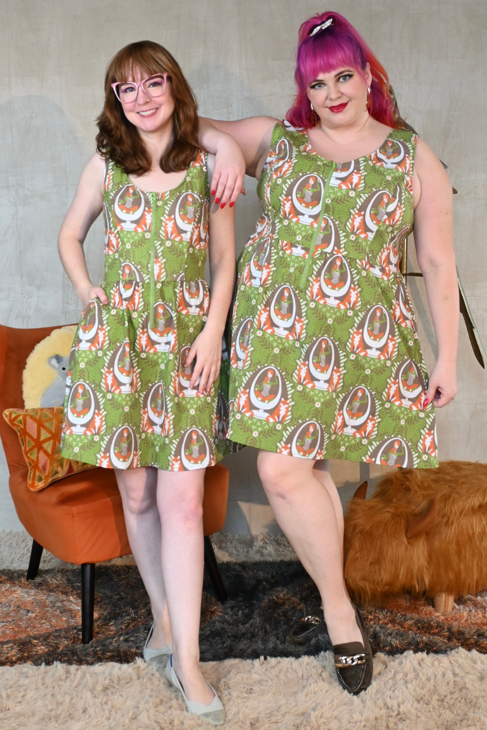 Two smiling models wearing olive green, orange, and brown fox and girl print dresses in an earth-toned room