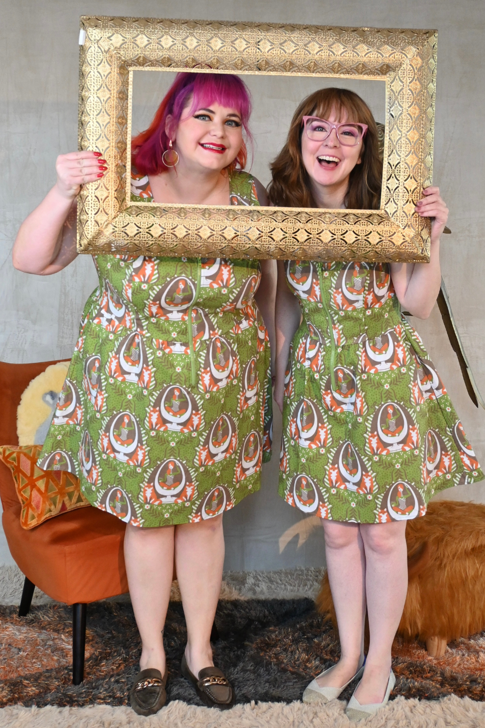 Two smiling models wearing matching green printed dresses, posing with gold picture frame