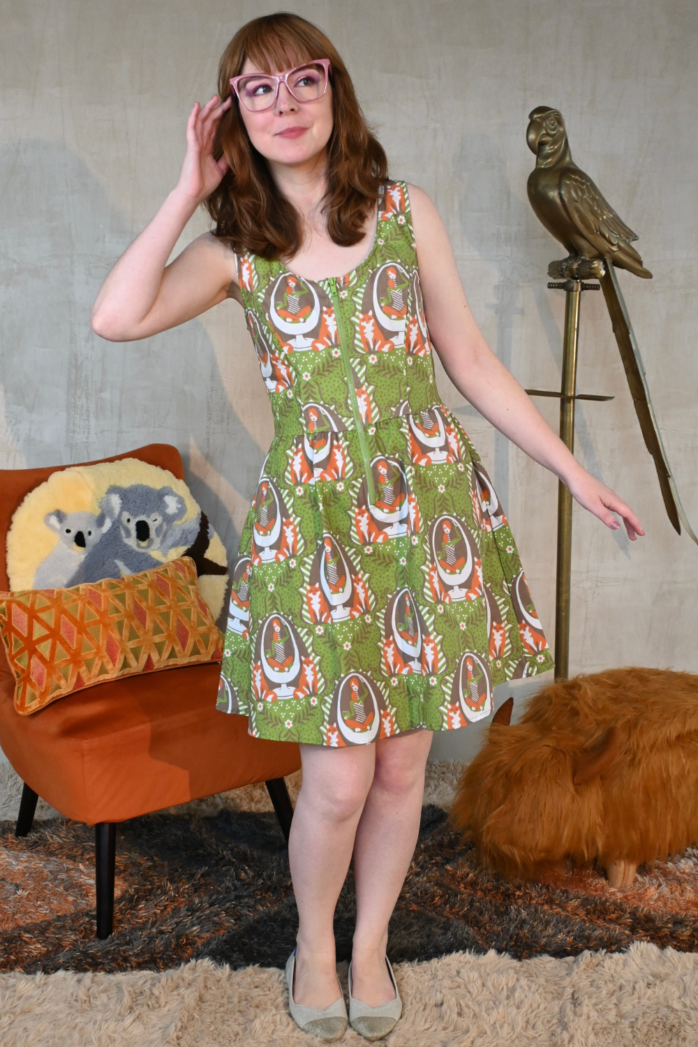 Cute brown-haired model wearing pink glasses and a zip-front olive green dress with orange, white and brown fox and egg chair print