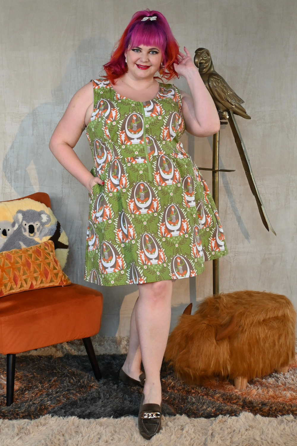 Cute pink-haired  plus size model wearing zip-front olive green dress with orange, white and brown fox and egg chair print