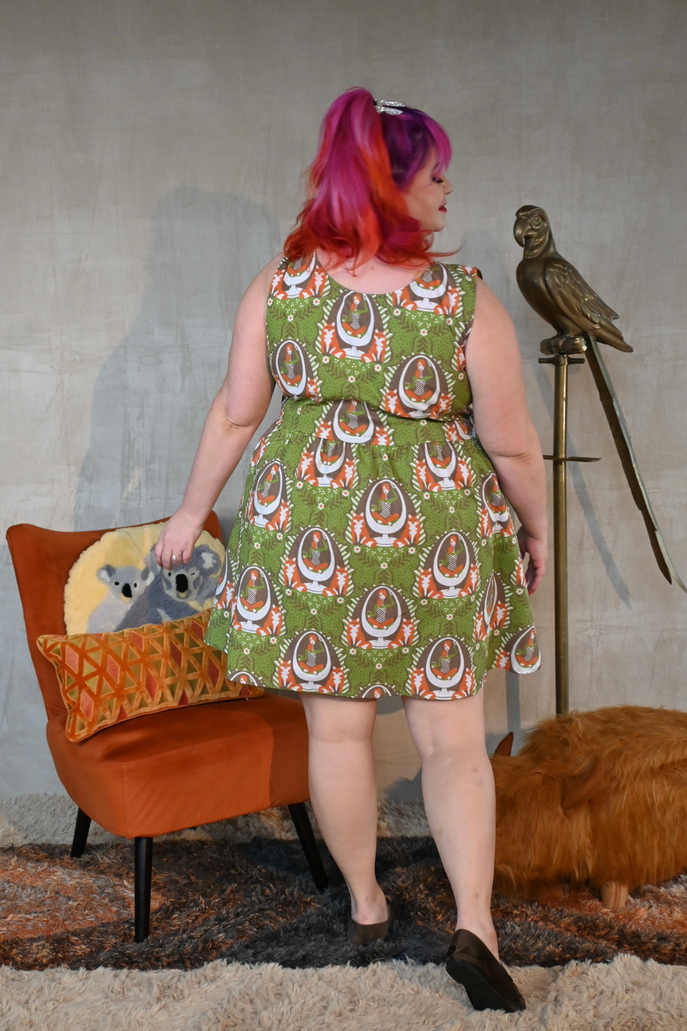 Cute pink-haired model wearing fit & flare olive green dress with orange, white and brown fox and egg chair print, back view