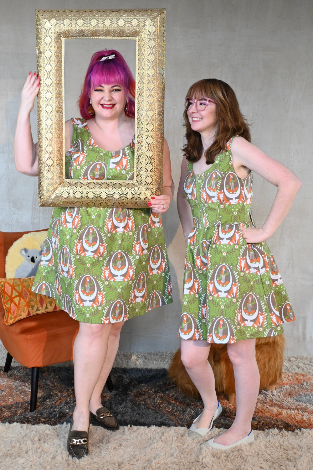 Cute pink-haired  plus size model and laughing brown-haired model wearing  a zip-front olive green dress with orange, white and brown fox and egg chair print