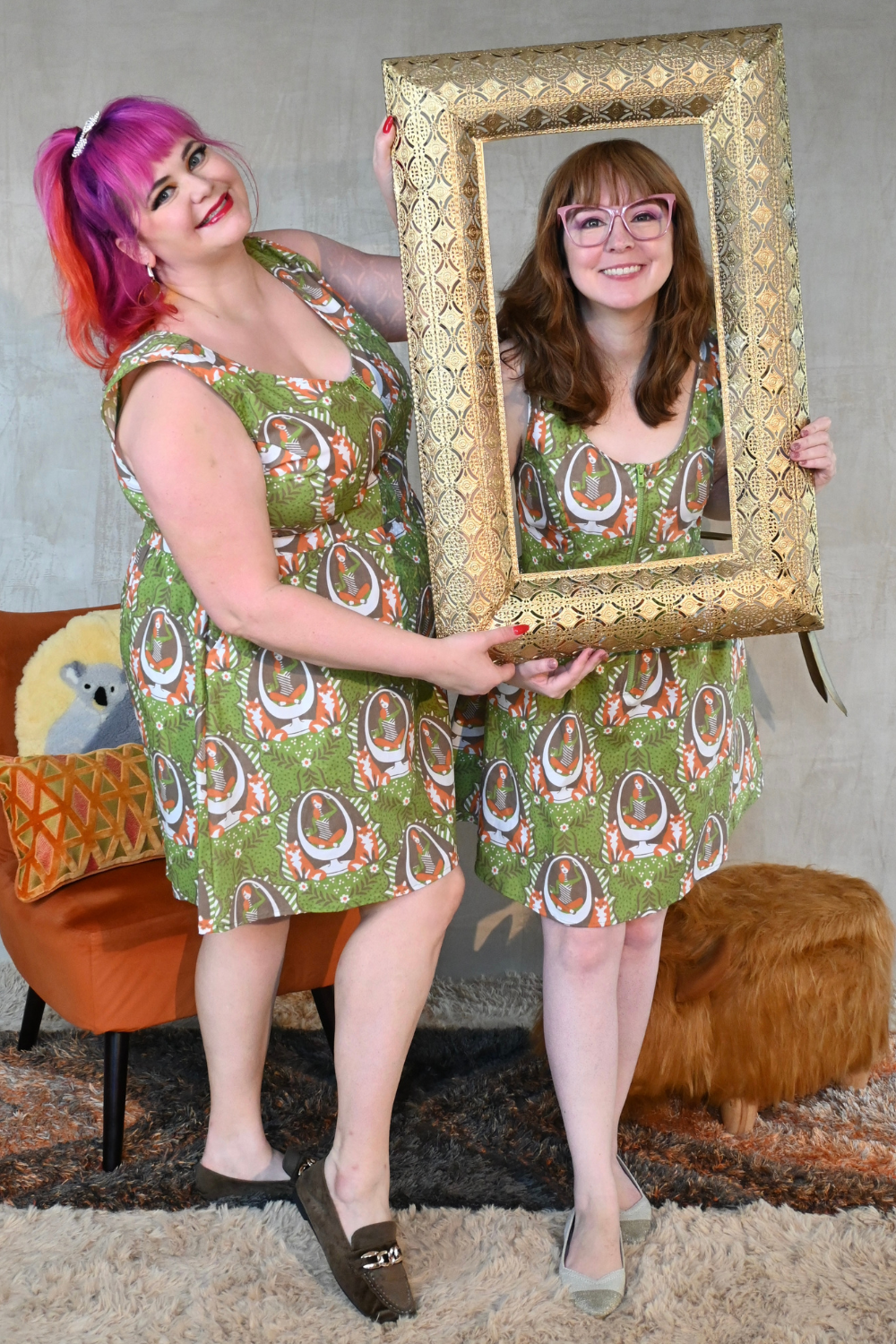 Two models wearing matching olive green fox and girl print scoopneck sleeveless dresses & posing with picture frame