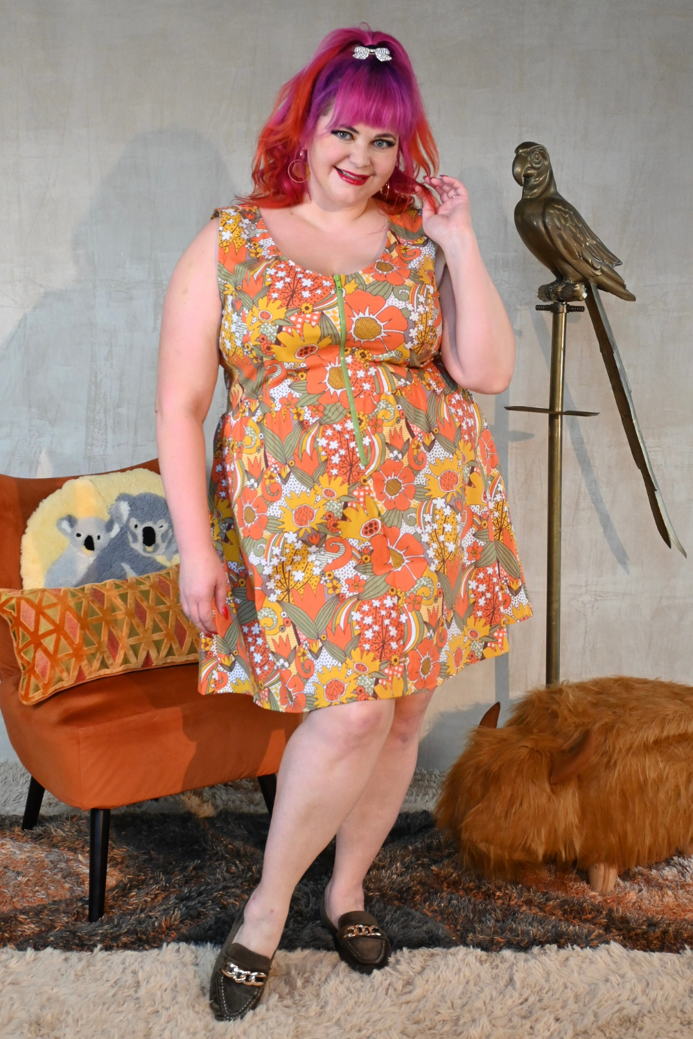 Pink haired curvy model wearing bold floral print knee-length dress, loafers in a living room