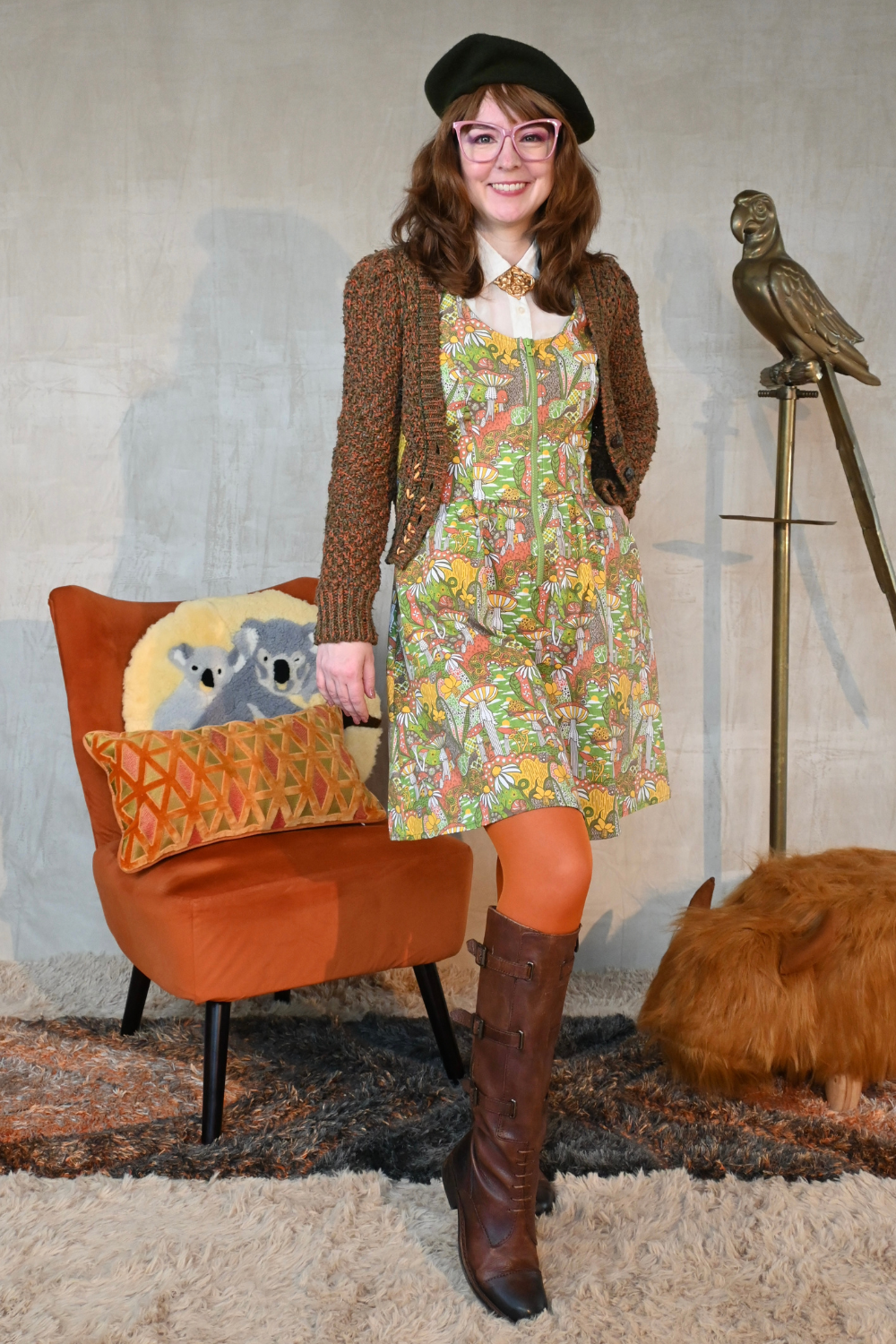 Cute brown-haired girl wearing mushroom print layered with cute cardigan, tights, boots and a beret in living room with bird statue